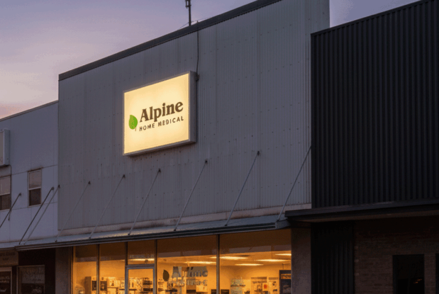 Exterior of Richfield Alpine Home Medical store at dusk, with the glowing green leaf logo sign and interior lights visible through the windows, reflecting a vibrant sunset on the street.
